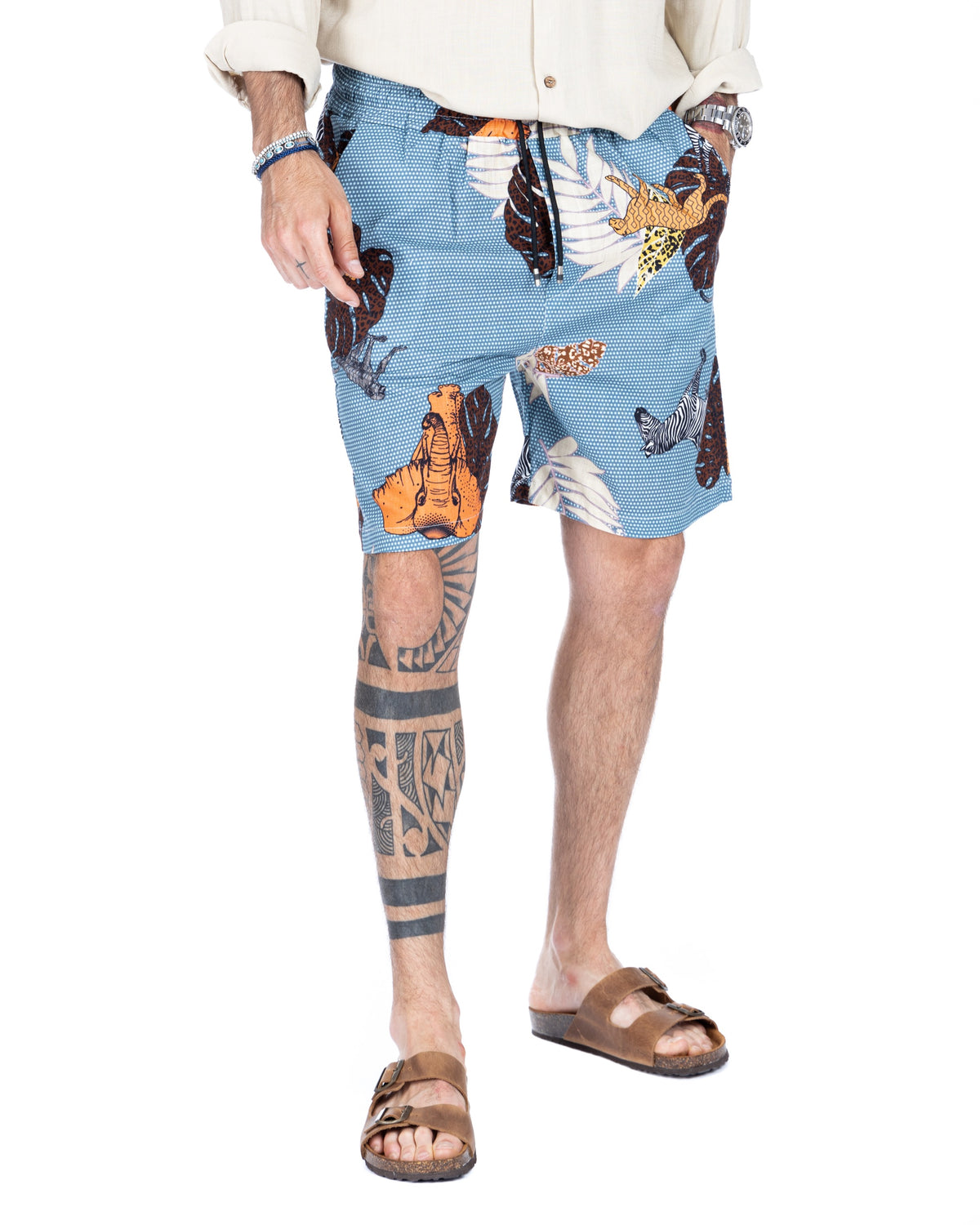 Men's light blue drawstring shorts with colorful safari animal print, styled with a cream shirt and slide sandals